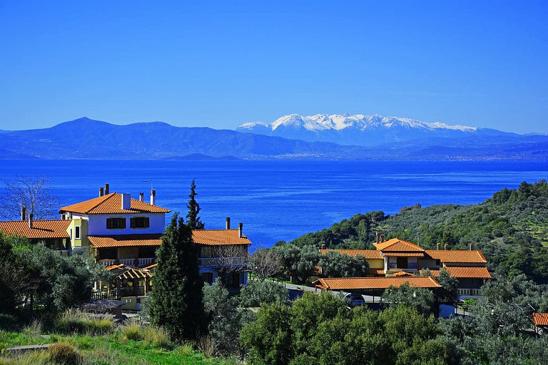 view from far of 'Vateri Guest House' complex buildings surrounded by trees and the sea in the background