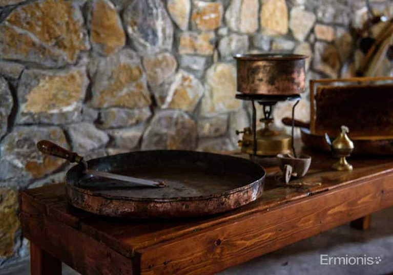 copper platter with spatula and other utensils on a table in front of stone wall at 'Honey Museum, Ermionis Bairaktaris Apiary'